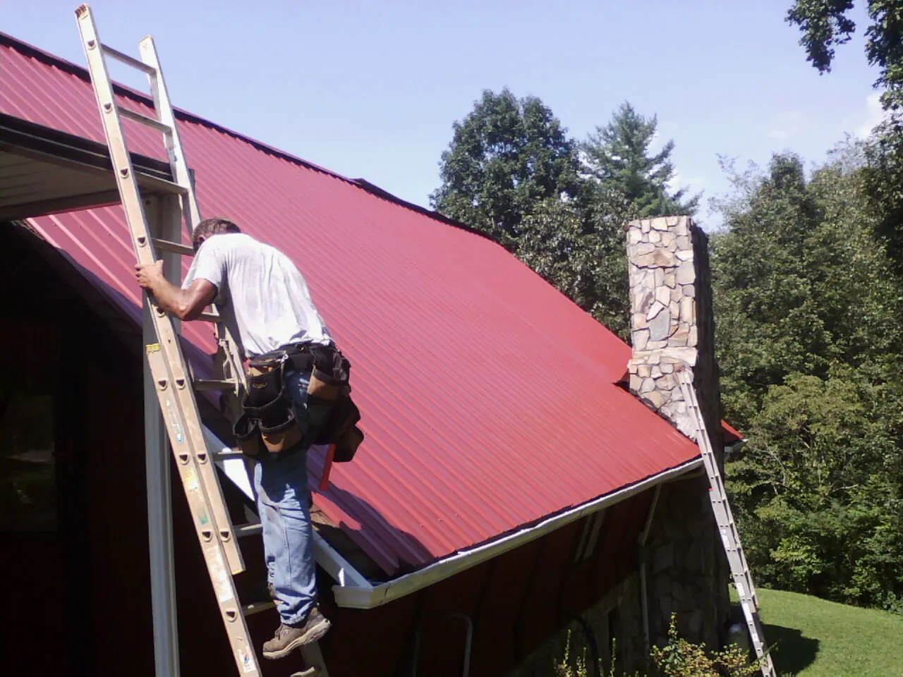 Roofer on a ladder beside a red metal roof and stone chimney — Soffit & Fascia Repair by Roofing Brothers in Polaris